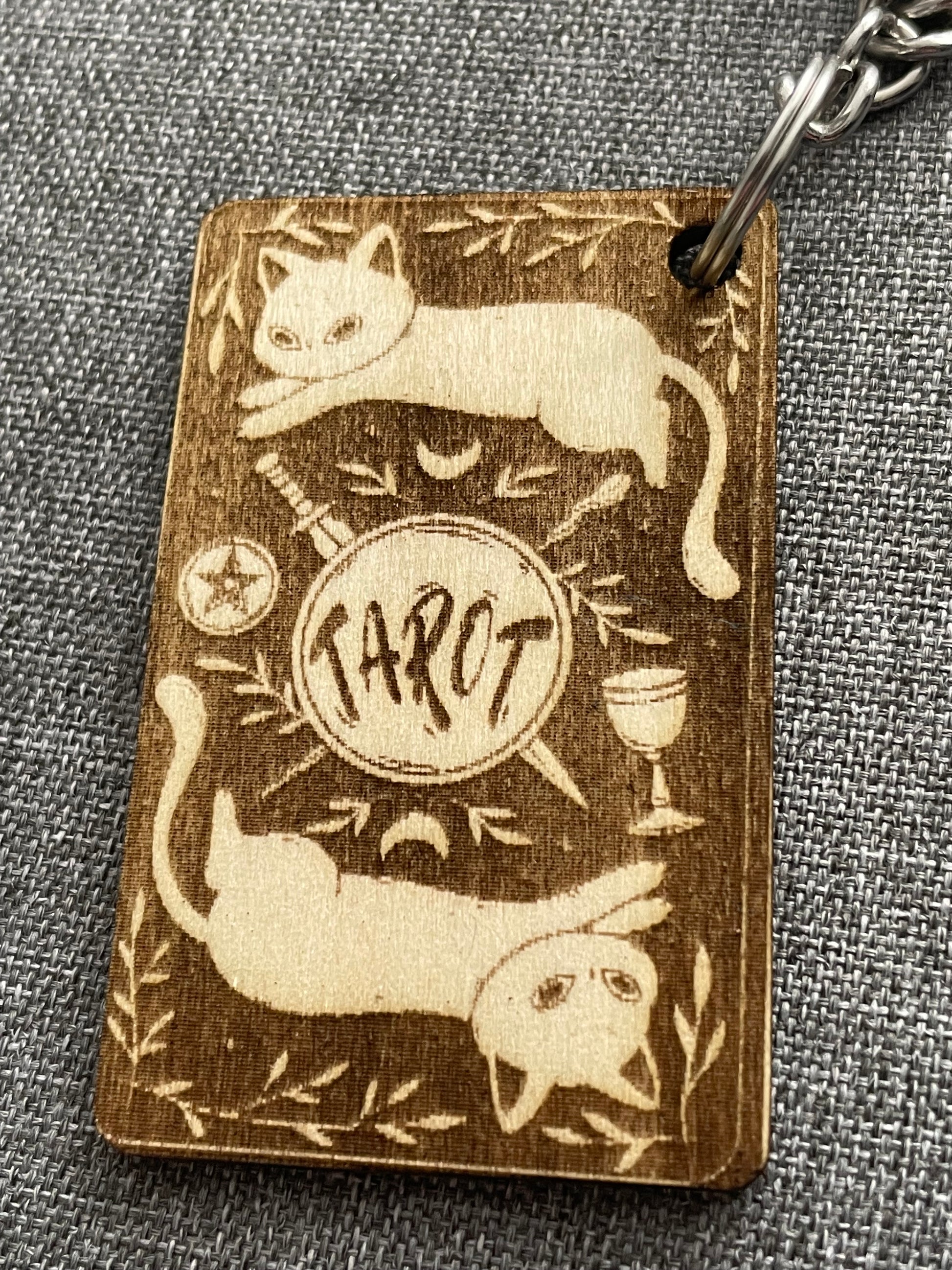 Wooden keychain with cat illustrations and a central emblem on a gray fabric background