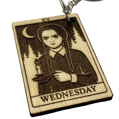 Wooden keychain with a design of a person holding a gun, labeled 'Wednesday', on a light gray background.